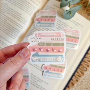 May include: A hand holds a stack of stickers featuring illustrated books. The stickers have a pink, blue, and green color scheme with the words "Recipes" and "Spring" on them. The stickers are on top of an open book.