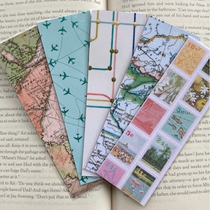 May include: A collection of five colorful bookmarks with different designs. The bookmarks feature maps, airplanes, flowers, and a flamingo. The bookmarks are placed on top of an open book.
