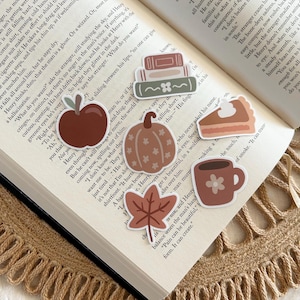 May include: A collection of fall-themed stickers, including an apple, pumpkin, leaf, pie slice, coffee mug, and books, arranged on an open book. The stickers are in shades of brown, orange, and green.