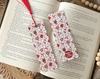 May include: Two Christmas-themed bookmarks with red tassels, lying on an open book. The bookmarks feature a repeating pattern of gingerbread houses, Santa hats, and bows. One bookmark has the text "mark it like it's hot".