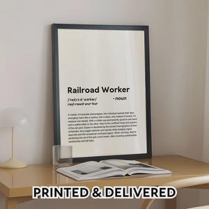 May include: A framed print with the text "Railroad Worker" and its phonetic pronunciation. The definition describes the role with humour. The print is on a desk with a glass and an open book. The words "PRINTED & DELIVERED" are at the bottom.