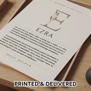 May include: A framed print featuring the name "Ezra" with a detailed explanation of its meaning and history. The print includes the text "HELP, HELPER" and "PRINTED & DELIVERED". The background is a light beige color, with a wooden frame.