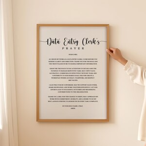May include: A framed wall art print featuring the text Data Entry Clerk's Prayer in black serif and script fonts on a light cream background. The prayer includes lines about focus, accuracy, and teamwork, enclosed in a simple light wood frame.