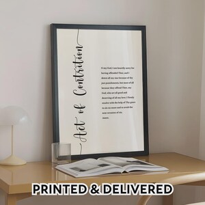May include: A framed wall art print featuring the Act of Contrition prayer in black text on a cream background. The title is written vertically in a script font, followed by the prayer text in a standard serif font, all enclosed in a black frame.