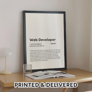Puede incluir: Una impresión enmarcada con el texto "Web Developer" y una definición. La impresión está en un marco negro y se encuentra sobre un escritorio de color claro con un libro y un vaso. Las palabras "PRINTED & DELIVERED" están en la parte inferior.