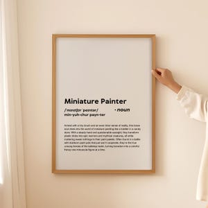 May include: A framed print with a white background and black text. The text defines "miniature painter" as a noun and provides a whimsical description of the hobby. The print has a wooden frame.