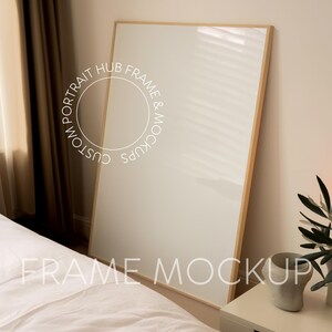 May include: A large, light wood frame mockup leaning against a white wall and a white bed. The frame is empty and ready for your design.