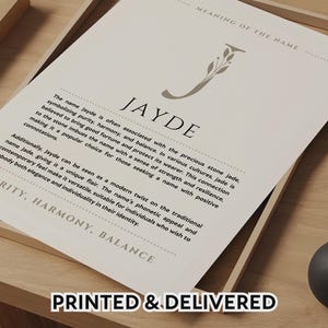 May include: A printed name meaning art print featuring the name "Jayde" in a serif font, with text describing the name's meaning and associations. The print is in a wooden frame, with the words "Printed & Delivered" at the bottom.