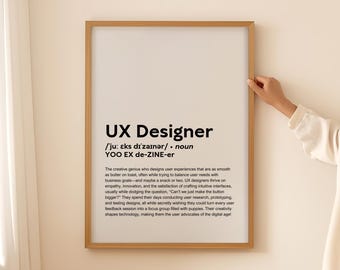May include: A framed print with the text "UX Designer" in a black font. The definition of a UX Designer is written below the title. The frame is a light brown color, and a person's hand is holding the frame.