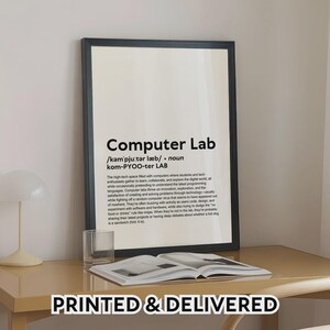 Puede incluir: Un cartel enmarcado con el texto "Computer Lab" en un marco negro. El cartel incluye una definición y descripción de un laboratorio de informática. Las palabras "PRINTED & DELIVERED" están en la parte inferior del marco. El cartel está sobre un escritorio con un vaso y un libro abierto.