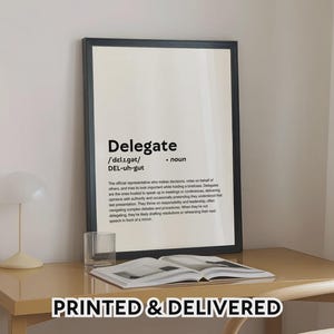 May include: A framed print with the definition of "Delegate" in black text on a cream background. The print is displayed on a light brown desk with a glass and an open book. The text "PRINTED & DELIVERED" is at the bottom.