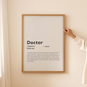 Puede incluir: Un cartel enmarcado con la definición de "Doctor" en texto negro. La definición incluye la pronunciación fonética y una descripción humorística del papel de un médico. El marco es de color marrón claro.