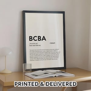 Pode incluir: Uma impressão emoldurada com o texto "BCBA" e uma definição. A impressão está em uma mesa com um copo e um livro aberto. As palavras "PRINTED & DELIVERED" estão na parte inferior.
