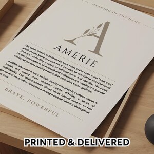 Puede incluir: Impresión enmarcada con el nombre "AMERIE" en letras grandes, acompañada de texto que describe su significado y origen. La impresión está en un marco de madera, con las palabras "PRINTED & DELIVERED" en la parte inferior.