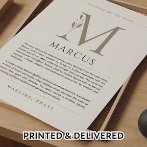 May include: A white paper print featuring the name Marcus in large serif font with a decorative leaf element. The text includes the heading Meaning of the Name, the name Marcus, a paragraph detailing its historical roots and significance, and the words Warlike, Brave at the bottom. The print is displayed in a light wood frame.