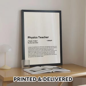 May include: A framed print with the text "Physics Teacher" in black font. The definition of a physics teacher is below the title. The print is displayed on a light-colored desk with a glass and an open book.