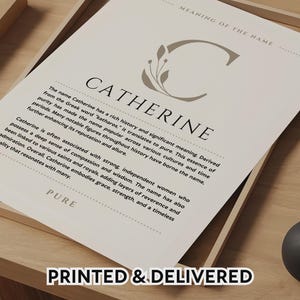 Puede incluir: Una impresión enmarcada con el nombre "Catherine" en letras grandes, con una "C" decorativa y texto que describe el significado y la historia del nombre. La impresión está en un marco de madera, con las palabras "PRINTED & DELIVERED" en la parte inferior.