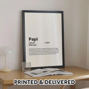 May include: A framed print with the definition of "Papi" in black text on a white background. The definition includes the pronunciation and a description of a loving father or grandfather. The phrase "PRINTED & DELIVERED" is at the bottom.