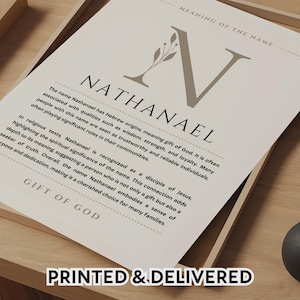 以下が含まれることがあります： スタイリッシュなフォントで「Nathanael」という名前が書かれた額入りのプリント。装飾的な「N」と、名前の意味と起源を説明するテキストが特徴です。プリントは木製のトレイに入っており、「PRINTED & DELIVERED」というテキストが含まれています。