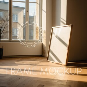 May include: A wooden frame mockup with a white background, sitting on a light wood floor with a herringbone pattern. The frame is leaning against a wall with a window in the background. Sunlight streams through the window, casting shadows on the floor and frame.
