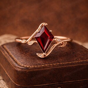 Peut inclure: Bague en or rose ornée d'une pierre rouge foncé en forme de cerf-volant. La monture de la bague présente un motif délicat en forme de vigne avec de petites pierres d'accent claires. La bague est posée sur un écrin en cuir marron.