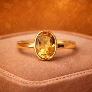 May include: A gold ring featuring an oval, yellow gemstone. The ring's band and setting are gold, and the gemstone is a warm, amber colour. The ring is displayed on a brown surface.