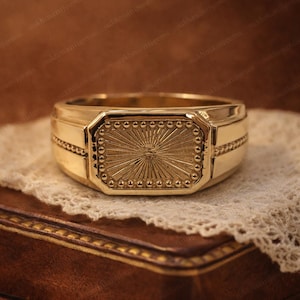 May include: A gold ring with a rectangular face, showcasing a sunburst pattern. The ring has a substantial band with decorative detailing and a polished finish. The ring is displayed on a white lace cloth.