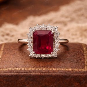 Pode incluir: Um anel de prata com uma marcante gema retangular vermelho-escuro, rodeada por um halo de pedras menores e transparentes. O anel é exibido sobre uma superfície castanha, realçando o design e o brilho da joia.
