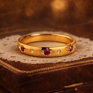 May include: A gold ring with three ruby gemstones and two small diamonds. The ring is set on a decorative lace doily atop a brown leather box. The ring's design features a band with evenly spaced gemstones.