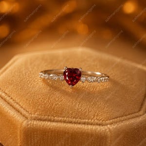 May include: A silver-toned ring featuring a central heart-shaped red gemstone. The band is accented with small, clear, marquise-cut stones on either side of the heart, creating a delicate and symmetrical design with a polished metallic finish.