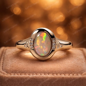 Könnte beinhalten: Ein goldener Ring mit einem ovalen Opal-Edelstein. Der Opal zeigt ein Farbspiel mit schillernden Farbtönen. Kleine Diamanten sind auf beiden Seiten des Opals gesetzt. Der Ring wird auf einer braunen Samtoberfläche präsentiert.