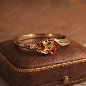 Puede incluir: Un anillo de oro con una gema de color amarillo-naranja en forma de pera. La banda del anillo tiene un diseño fluido, que sostiene la piedra. El anillo se exhibe en un joyero de cuero marrón, resaltando su elegante diseño.