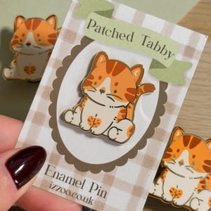 May include: Enamel pin featuring a cartoon orange tabby cat with white accents. The pin is on a card that says "Patched Tabby" and "Enamel Pin izzoo.co.uk". Additional pins are visible in the background.
