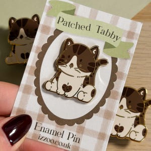 May include: Enamel pin featuring a brown and white tabby cat design with gold trim. The pin is on a card with the text "Patched Tabby" and "Enamel Pin izzoo.co.uk". Two additional pins are visible in the background.