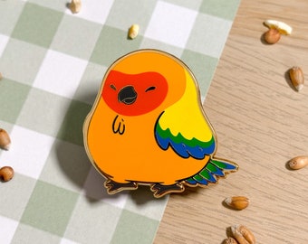 May include: A gold-colored enamel pin of a cartoon orange and yellow bird with a red head and blue and green wings. The bird is winking and has a small smile.
