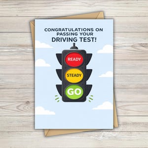 Peut inclure: Une carte de vœux sur fond bleu clair avec des nuages blancs et une illustration de feu de signalisation noir. Le texte indique CONGRATULATIONS ON PASSING YOUR DRIVING TEST! Le feu comporte des cercles rouge, jaune et vert marqués READY, STEADY, GO.