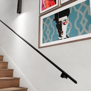 May include: A black metal handrail is mounted to a white wall. The handrail extends from the top of the image to the bottom right corner. A wooden staircase with brown steps is visible in the background. There are framed pictures on the wall above the handrail.