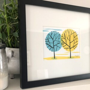 May include: A framed print of two stylized trees with black branches. The tree on the left has blue leaves and the tree on the right has yellow leaves. The print is titled "Two Trees" and signed "J.S. 2019".