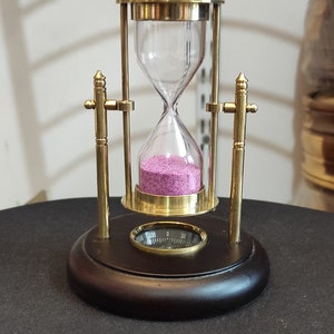 May include: A brass hourglass with a pink sand timer and a compass on a wooden base.