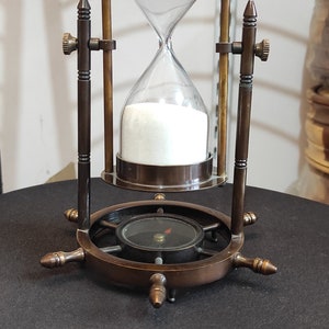 May include: A brass hourglass with a compass base. The hourglass is filled with white sand and has a clear glass bulb. The compass base is a ship's wheel with a compass needle pointing north.