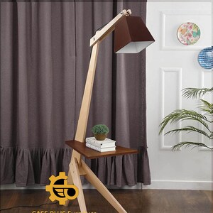 K&ouml;nnte beinhalten: Eine Stehlampe aus Holz mit einem braunen Lampenschirm und einem kleinen Regal. Die Lampe hat ein einzigartiges Design mit einem langen, abgewinkelten Arm und einem dreieckigen Sockel. Das Regal ist perfekt f&uuml;r ein Buch oder eine kleine Pflanze.