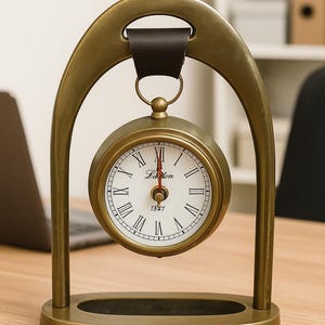 May include: A brass-toned metal table clock suspended from an arched frame by a dark brown leather strap. The clock face is white with black Roman numerals, black hands, a red second hand, and the text "Lalon" and "1847" printed in the center.