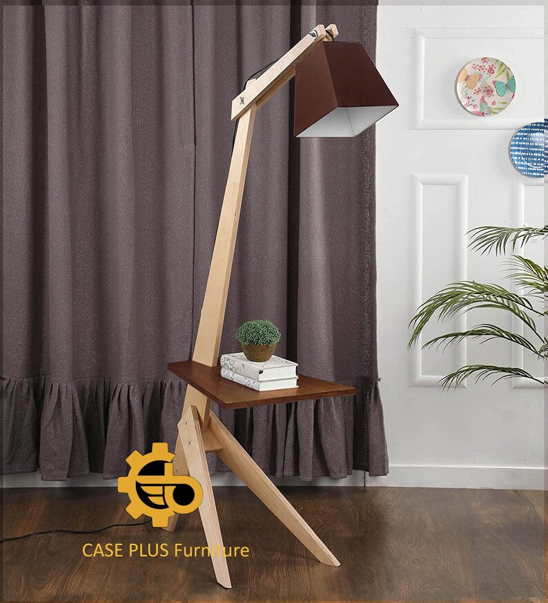 K&ouml;nnte beinhalten: Eine Stehlampe aus Holz mit einem braunen Lampenschirm und einem kleinen Regal. Die Lampe hat ein einzigartiges Design mit einem langen, abgewinkelten Arm und einem dreieckigen Sockel. Das Regal ist perfekt f&uuml;r ein Buch oder eine kleine Pflanze.
