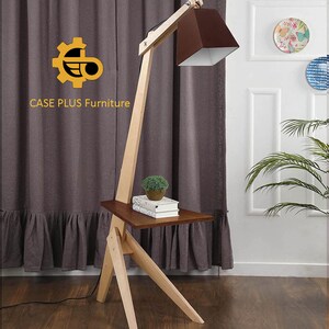 K&ouml;nnte beinhalten: Eine Stehlampe aus Holz mit einem braunen Lampenschirm und einem kleinen Regal. Die Lampe hat einen langen, verstellbaren Arm und einen Holzfu&szlig;. Das Regal ist aus Holz und hat eine braune Oberfl&auml;che. Die Lampe steht in einem Raum mit einem braunen Vorhang und einer wei&szlig;en Wand.