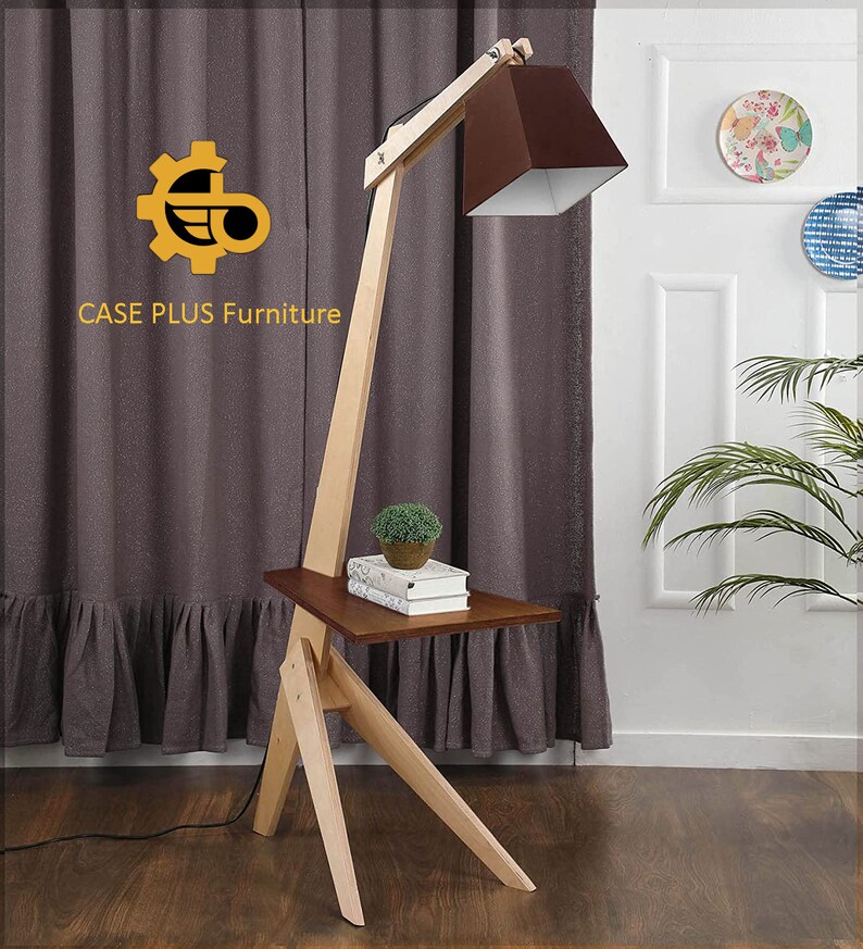 K&ouml;nnte beinhalten: Eine Stehlampe aus Holz mit einem braunen Lampenschirm und einem kleinen Regal. Die Lampe hat einen langen, verstellbaren Arm und einen Holzfu&szlig;. Das Regal ist aus Holz und hat eine braune Oberfl&auml;che. Die Lampe steht in einem Raum mit einem braunen Vorhang und einer wei&szlig;en Wand.