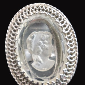 May include: A silver oval-shaped brooch with a clear glass cameo featuring a woman's profile.
