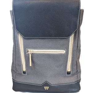 May include: Gray backpack with a black flap and base, featuring gold zippers and white stitching. The flap and base are made of a textured material. A gold emblem is visible on the bottom.