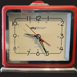 May include: A red alarm clock with a white face and black hands. The clock face has black numbers and the text "skytimer" in black. The clock is set to 7:55.