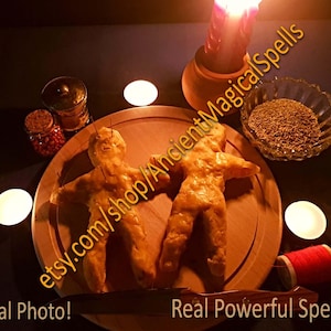 Può includere: Due bambole voodoo fatte a mano su un piatto di legno, circondate da candele, spezie e una bobina di filo rosso. Sono presenti anche una candela accesa e una piccola ciotola di vetro piena di erbe. Il testo "Real Photo!" e "Real Powerful Spell!" sono visibili.
