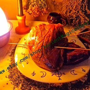 May include: A raw, reddish-brown heart impaled with wooden skewers, presented on a yellow plate adorned with crescent moon and symbol designs. The plate is surrounded by dried herbs and incense cones, with a candle and cup in the background.